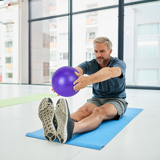 "Man performing seated Pilates exercise with purple exercise ball on blue mat by window"
