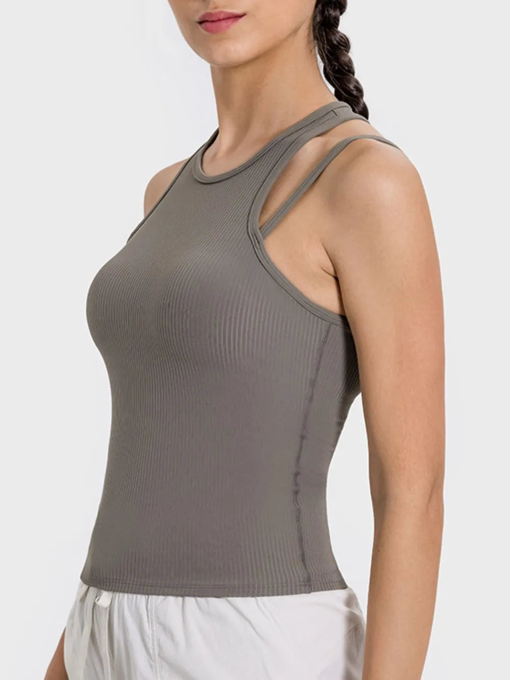 "Woman wearing minimalist taupe workout top with distinctive double straps and ribbed fabric, paired with white pants."
