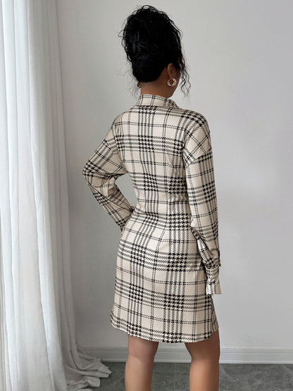 Person wearing a plaid dress standing against a white wall with curtains.